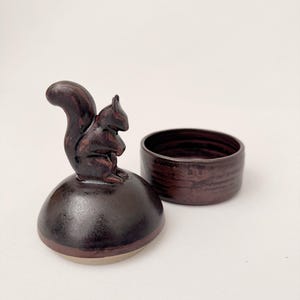 Puede incluir: Una caja de baratijas de cerámica con acabado marrón oscuro. La tapa presenta una detallada figura de ardilla. La caja redonda es de color marrón oscuro a juego. La base de la caja es de color beige claro.