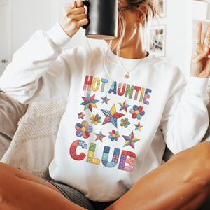 May include: White sweatshirt with the text "HOT AUNTIE CLUB" in colorful letters and star and flower graphics. The person in the image is holding a black mug.