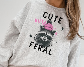 May include: Gray sweatshirt with a raccoon wearing a pink hat and the text "Cute but Feral" printed on it.