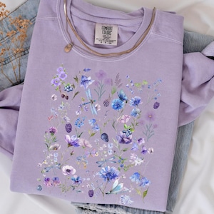 May include: A lavender sweatshirt with a floral print featuring blue, purple, and white flowers. The print includes dragonflies and small berries. The sweatshirt is laid flat on a white surface.
