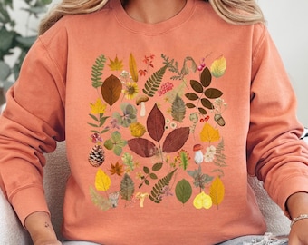 May include: Terracotta colored sweatshirt with a colorful print of leaves, flowers, and other botanical elements. The print is centered on the front of the sweatshirt.