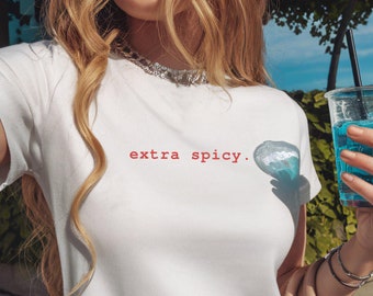May include: White crop top with the text "extra spicy" in red. The shirt has short sleeves and a crew neck. The image also shows a clear plastic cup with a blue drink and a brown purse.