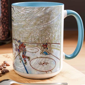 Puede incluir: Una taza grande de cer&aacute;mica blanca con interior y asa azul claro. La taza presenta una ilustraci&oacute;n detallada de un partido de hockey en curso, con jugadores, un portero y una multitud en el fondo. El dise&ntilde;o es de estilo art&iacute;stico y detallado.
