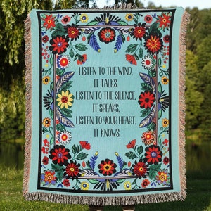 May include: A turquoise throw blanket with a floral border and the text "LISTEN TO THE WIND, IT TALKS. LISTEN TO THE SILENCE, IT SPEAKS. LISTEN TO YOUR HEART, IT KNOWS." The blanket has a fringe edge and is held outdoors.