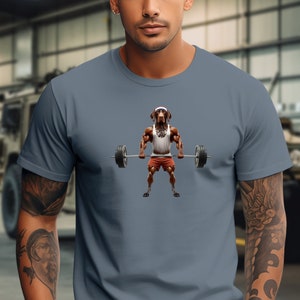 May include: A steel blue t-shirt featuring a cartoon image of a dog wearing a Santa hat and lifting weights. The dog is wearing red shorts and a white tank top.