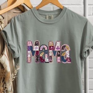 May include: A sage green t-shirt displaying the word "Mama" in a patchwork design. The letters are made up of various patterns and colours, including purple, pink, and blue. The shirt is hanging on a wooden hanger.