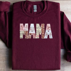 May include: A maroon sweatshirt featuring the word "MAMA" in a decorative font. The letters are filled with a collage of floral and musical note patterns. The sweatshirt is displayed on a wooden surface.