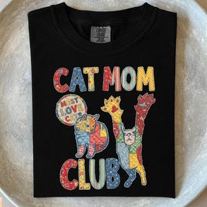 May include: Black crew neck t-shirt with the text "CAT MOM CLUB" in colorful patchwork letters. The shirt features a patchwork design of two cats and a circle that says "MUST LOVE CATS".