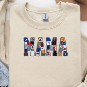 May include: Sand-colored sweatshirt with the word "MAMA" embroidered in a patchwork design. The letters are composed of various fabric patterns, including denim, plaid, and star motifs. The sweatshirt has a crew neck and long sleeves.