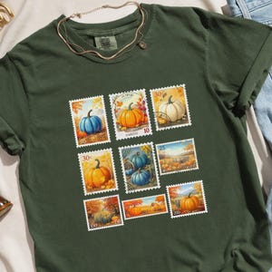 以下が含まれることがあります： モスグリーンのTシャツ。水彩画の様々な色のカボチャを描いた切手のデザインが特徴です。 シャツの袖はロールアップされており、下部には「Moss」の文字がプリントされています。