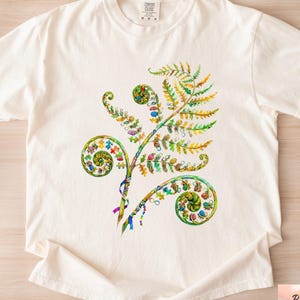 May include: Ivory-colored t-shirt featuring a colorful fern design. The fern has green, yellow, and blue accents, with spiral-shaped fronds. The shirt has a crew neck and short sleeves. The text "Ivory" is at the bottom left.