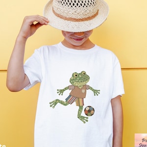 May include: White t-shirt featuring a cartoon frog playing soccer. The frog is wearing a brown shirt and striped shorts. The image also includes a straw hat. The text "white" is at the bottom left.