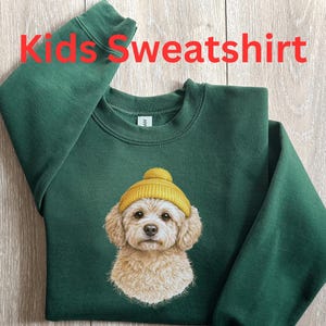 May include: Forest green kids' sweatshirt featuring a graphic of a dog wearing a yellow beanie. The text "Kids Sweatshirt" is printed in red at the top. The sweatshirt is made of a soft material.