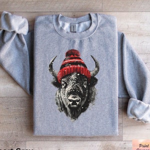 May include: A gray sweatshirt with a graphic of a bison wearing a red and white winter hat. The bison is looking directly at the viewer. The sweatshirt is folded in half and laid flat on a wooden surface.