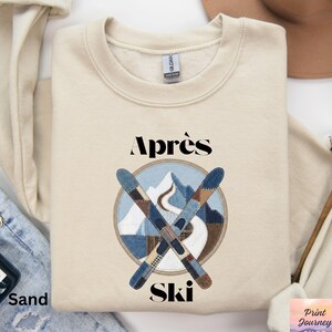 May include: A sand-colored sweatshirt with the words "Après Ski" in black. The design features crossed skis and a mountain scene in a circular patch. The sweatshirt is folded on a pair of blue jeans.
