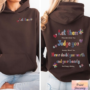 Peut inclure: Sweat à capuche marron chocolat foncé avec une poche avant et une capuche. Le devant du sweat à capuche porte le texte "Let them" brodé en fil coloré. Le dos du sweat à capuche porte le texte "Let them, Judge you, Never doubt your worth and your beauty, And Keep Shining."