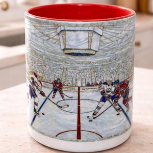 Puede incluir: Taza de cer&aacute;mica con interior rojo y exterior blanco. La taza presenta una ilustraci&oacute;n detallada de un partido de hockey en curso, con jugadores con uniformes azules y rojos. El dise&ntilde;o incluye una multitud y el estadio.