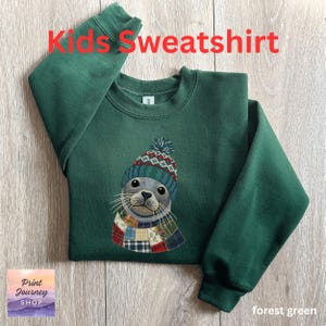 May include: Forest green kids' sweatshirt featuring a seal wearing a patterned hat and scarf. The text "Kids Sweatshirt" is displayed in red at the top. The sweatshirt has a crew neck and long sleeves. The Print Journey Shop logo is in the bottom left corner.
