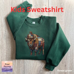 May include: Forest green kids' sweatshirt with the text "Kids Sweatshirt" in red. The sweatshirt features an embroidered bison wearing a plaid hat and patchwork design. The sweatshirt is a crew neck style.