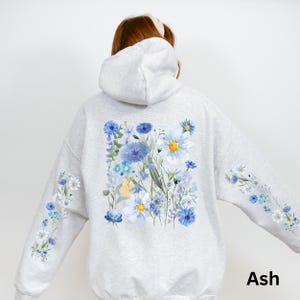 May include: Light gray hoodie with a floral design. The back of the hoodie features a large watercolor-style arrangement of blue, white, and yellow flowers. The sleeves have smaller floral patterns. The word "Ash" is printed on the lower right.