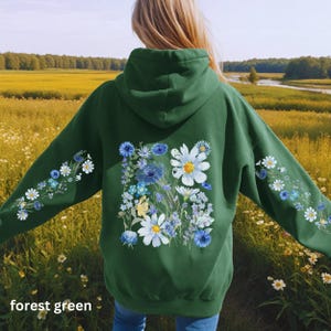 May include: Forest green hoodie with a floral design. The back of the hoodie features a large arrangement of white daisies, blue cornflowers, and other wildflowers. The sleeves have smaller floral patterns. The text "forest green" is at the bottom.