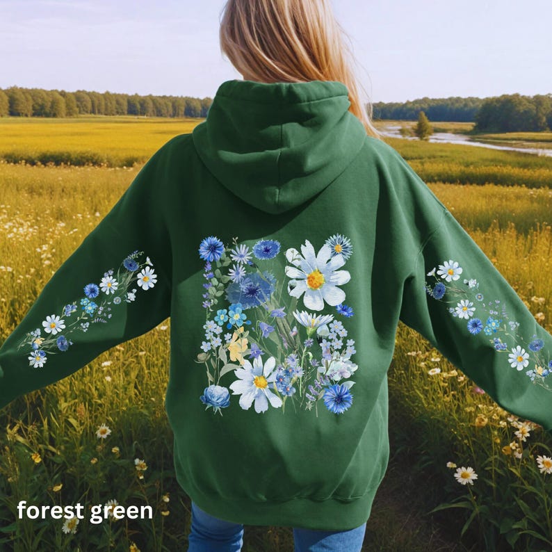 May include: Forest green hoodie with a floral design. The back of the hoodie features a large arrangement of white daisies, blue cornflowers, and other wildflowers. The sleeves have smaller floral patterns. The text "forest green" is at the bottom.