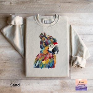 May include: Sand-colored sweatshirt featuring a colorful parrot wearing a knitted hat. The parrot has vibrant blue, yellow, red, and green feathers. The sweatshirt has long sleeves and a crew neck. The text "Print Journey Shop" is in the bottom right corner.