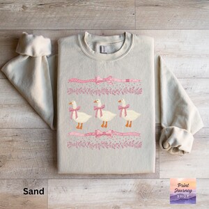 May include: Sand-colored sweatshirt featuring an embroidered design of three geese wearing pink bows. Above and below the geese are pink ribbons with bows and floral accents. The sweatshirt has long sleeves and a crew neck.