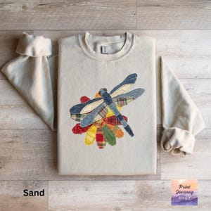 May include: A sand-coloured sweatshirt with a patchwork dragonfly design. The dragonfly has blue denim and plaid wings, with a body of blue and yellow fabric. The dragonfly is perched on a flower made of colourful patchwork. The text "Print Journey Shop" is in the lower right corner.