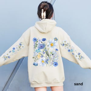 May include: Sand-colored hoodie with a floral design. The back features a large square of blue and white flowers, with smaller floral patterns on the sleeves. The word "sand" is printed at the bottom.