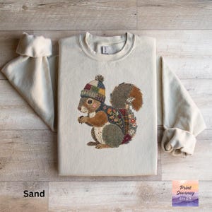 May include: Sand-colored sweatshirt featuring an embroidered squirrel wearing a colorful knit hat. The squirrel has a patchwork design with floral and plaid patterns. The sweatshirt has long sleeves with rolled cuffs. The text "Sand" is at the bottom left.