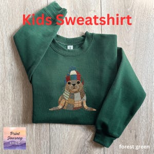 May include: Forest green kids' sweatshirt with the text "Kids Sweatshirt" in red. The sweatshirt features an embroidered seal wearing a colorful hat and scarf. The sweatshirt is made of a soft material.