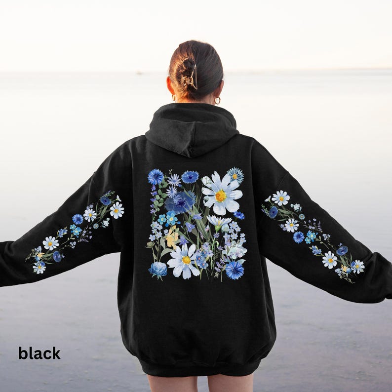 May include: Black hoodie with a floral design. The back of the hoodie features a large arrangement of white daisies and blue flowers. The sleeves have a smaller floral pattern. The word "black" is printed at the bottom.