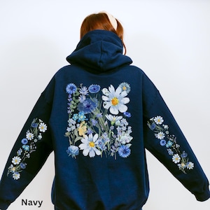 May include: Navy blue hoodie with a floral design. The back features a large arrangement of white and blue flowers. Floral patterns also adorn the sleeves. The word "Navy" is printed at the bottom.