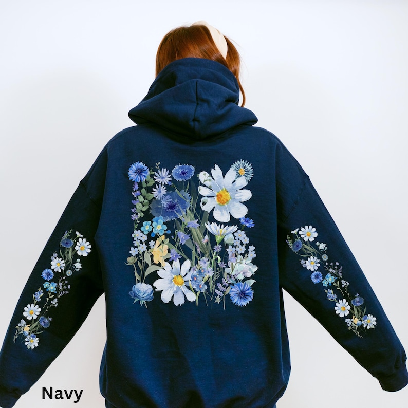 May include: Navy blue hoodie with a floral design. The back features a large arrangement of white and blue flowers. Floral patterns also adorn the sleeves. The word "Navy" is printed at the bottom.