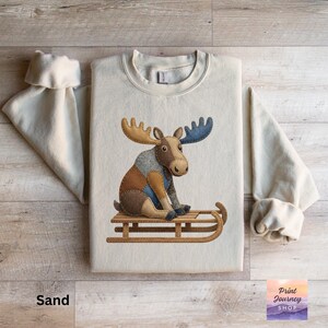 May include: A sand-colored sweatshirt featuring a whimsical moose illustration. The moose, with patchwork details in brown, gray, and blue, sits on a wooden sled. The text "Sand" is at the bottom left.