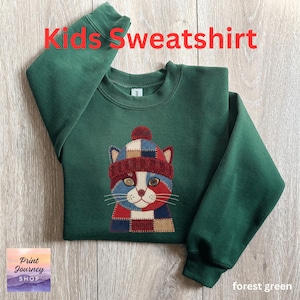 May include: Forest green kids' sweatshirt with the text "Kids Sweatshirt" in red. The sweatshirt features an embroidered cat wearing a patchwork hat. The cat's face is red, white, and blue. The sweatshirt is made of a soft material.