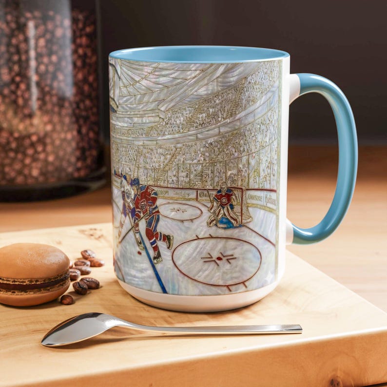 Puede incluir: Una taza grande de cer&aacute;mica blanca con interior y asa azul claro. La taza presenta una ilustraci&oacute;n detallada de un partido de hockey en curso, con jugadores, un portero y una multitud en el fondo. El dise&ntilde;o es de estilo art&iacute;stico y detallado.
