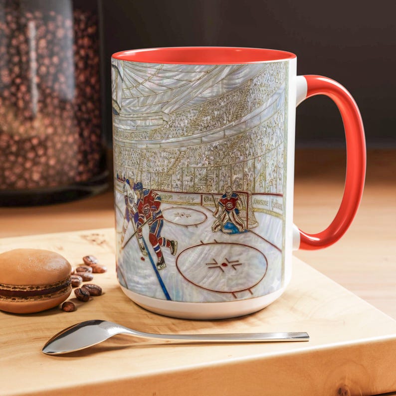 Puede incluir: Taza de cer&aacute;mica blanca con interior y asa rojos, con una ilustraci&oacute;n detallada de un partido de hockey. La obra de arte representa jugadores en el hielo, un portero y una multitud en las gradas. Una cuchara y un macaron est&aacute;n sobre la superficie de madera.