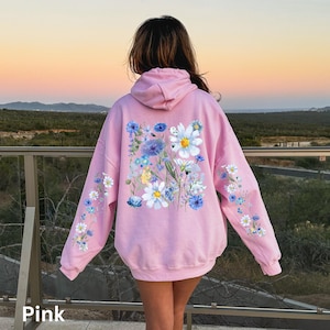 May include: A light pink hoodie with a floral design. The back of the hoodie features a large arrangement of white and blue flowers. Smaller floral patterns adorn the sleeves. The word "Pink" is at the bottom left.