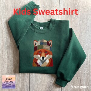 May include: Forest green kids' sweatshirt featuring an embroidered fox wearing a plaid hat. The text "Kids Sweatshirt" is in red at the top. The sweatshirt has a crew neck and long sleeves. The Print Journey Shop logo is in the bottom left corner.