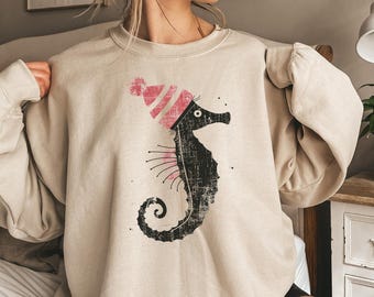 May include: A beige sweatshirt featuring a black and pink seahorse graphic. The seahorse is wearing a pink and white striped hat. The text "Gildan 18000 Sand" is printed on the bottom of the sweatshirt.