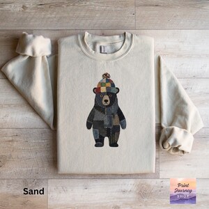 May include: Sand-colored sweatshirt featuring a bear illustration. The bear is depicted wearing a patchwork design and a colorful knit hat. The text "Print Journey Shop" is visible in the lower right corner. The word "Sand" is printed at the bottom left.