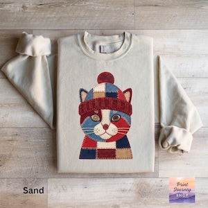 May include: A sand-colored sweatshirt features a whimsical cat design wearing a patchwork hat. The cat's face is composed of blue, red, and white patches, with button eyes and a red nose. The text "Print Journey Shop" is visible in the bottom right corner.