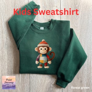 May include: Forest green kids' sweatshirt with a monkey graphic. The monkey wears a knit hat and patchwork clothing. The text "Kids Sweatshirt" is in red at the top. The shop logo is in the bottom left corner.