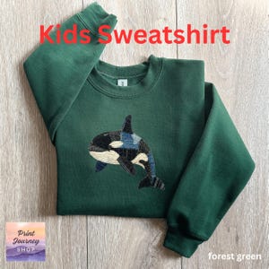 May include: A forest green children's sweatshirt featuring an orca whale design. The whale is made from black, white, and blue denim patches. The words "Kids Sweatshirt" are in red. The sweatshirt has a crew neck.