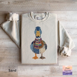 Peut inclure: Sweat-shirt couleur sable avec un motif de canard brodé. Le canard a une tête bleue, un bec jaune et un corps en patchwork avec une écharpe colorée. Le sweat-shirt a des manches longues et un col rond.