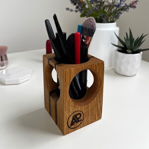 Könnte beinhalten: Ein quadratischer Kosmetikpinselhalter aus Holz mit abgerundeten Ausschnitten. Der Halter ist aus hellem braunem Holz mit einer dunkelbraunen Beize. Er hat ein schwarzes Logo auf der Unterseite, das "AP" lautet.
