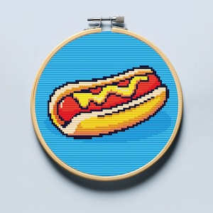 May include: A cross-stitch embroidery hoop with a pixelated image of a hot dog with mustard and ketchup on a blue background.