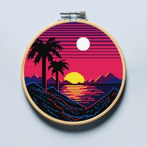 May include: A cross-stitch embroidery hoop with a pixelated sunset scene. The scene features two palm trees, a yellow sun setting over a blue body of water, and a white moon in the sky. The background is a gradient of pink, purple, and blue.